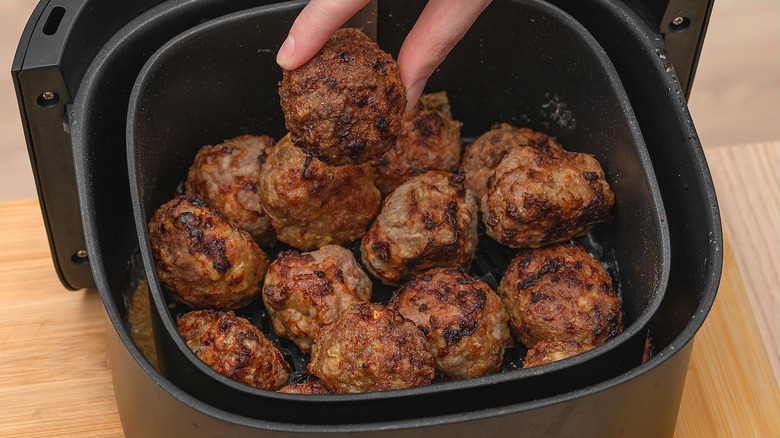 Two fingers holding a cooked meatball over an air fryer basket full of meatballs