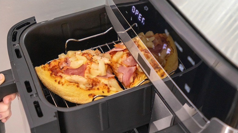 Hand holding an air fryer basket with two slices of pizza topped with pineapple, ham, and cheese