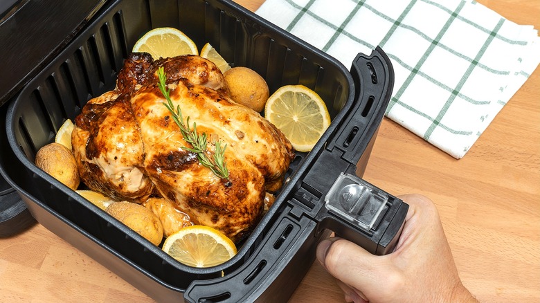 A hand holding an air fryer basket filled with a whole cooked chicken, baby potatoes, and lemon slices
