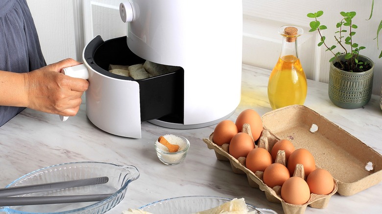 Hand pulling out air fryer basket next to a carton of brown eggs and two glass casserole dishes