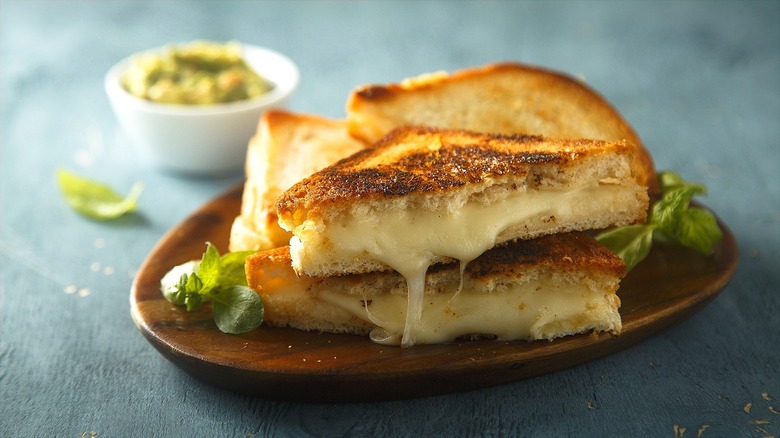Halved, melty grilled cheese sandwiches on a circular, wooden plate with fresh herbs