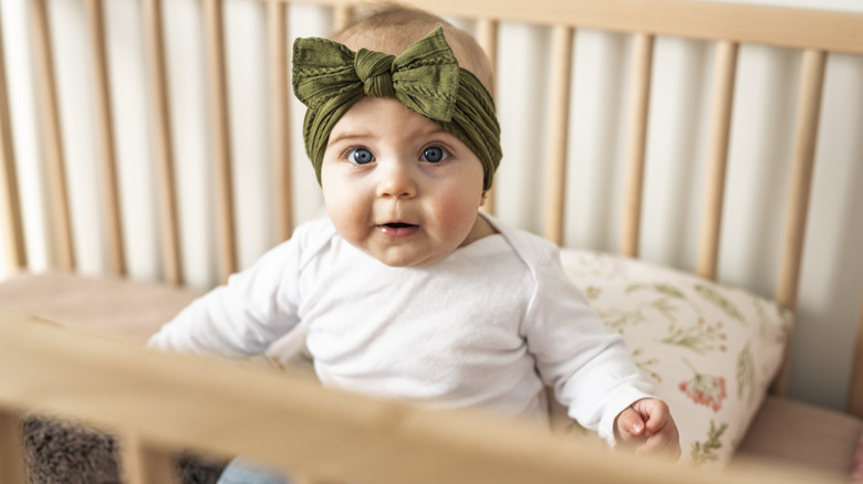 Baby girl with green hair bow standing in crib