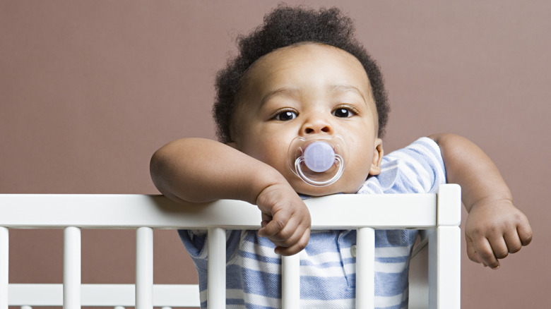 A baby boy stands up in his crib with a pacifier in his mouth and his arms hanging over the rails