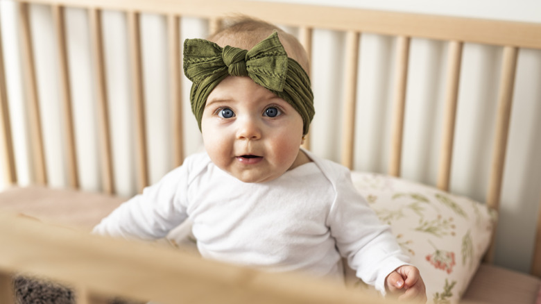 A baby girl wearing a bow headband sitting up in a crib