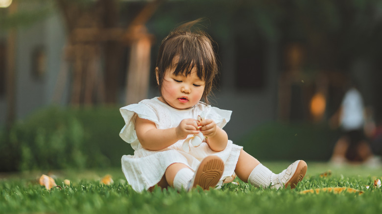 A toddler girl sits in the grass focused on something in her hands