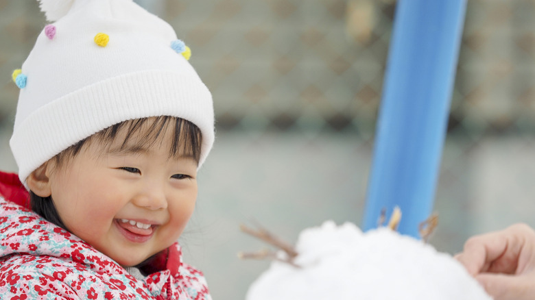 Toddler girl in white snow hat and jacket smiling at snowman