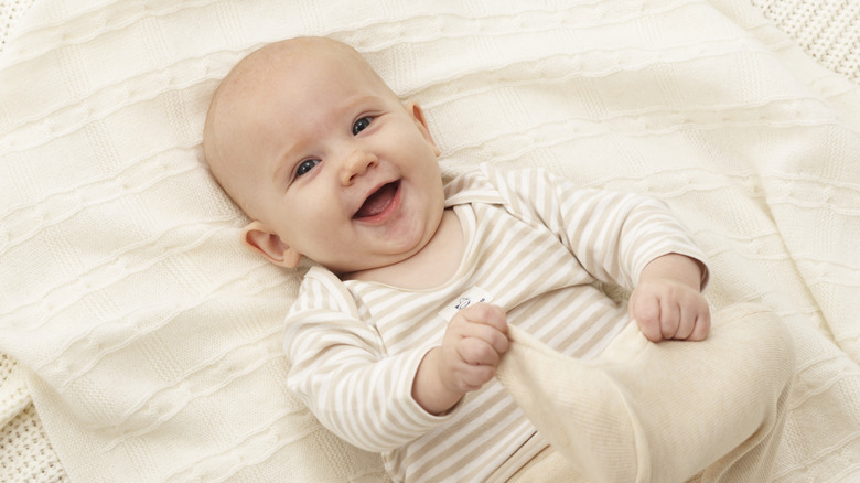 A baby lies on ivory linens smiling and holding his foot