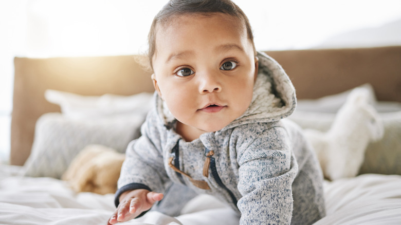 A baby crawls on a bed and looks straight at the camera