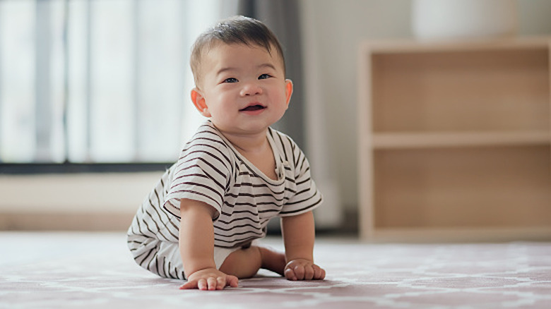 A baby sits up on the floor and smiles toward the camera