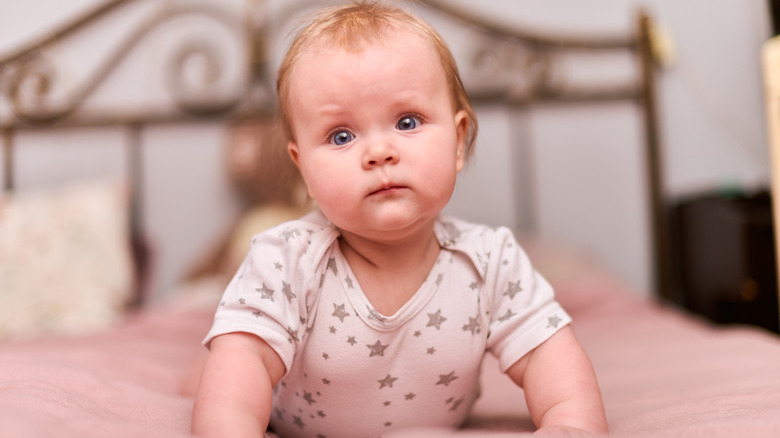 A baby girl wearing a onesie with stars holds herself up on her arms on a bed