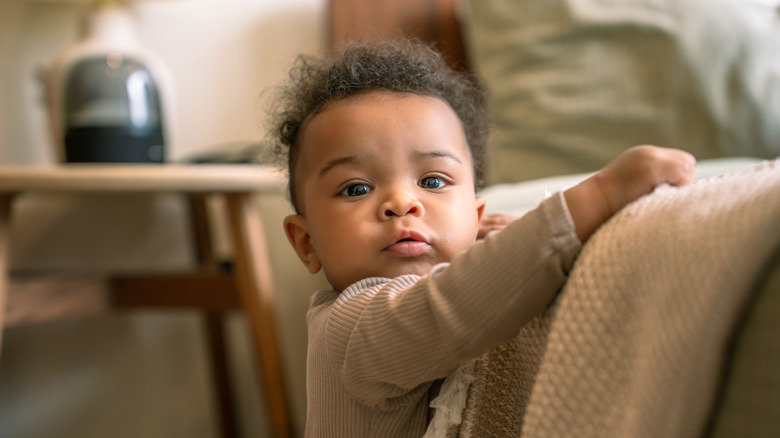 A baby boy holds himself up on the side of a couch, gripping a blanket