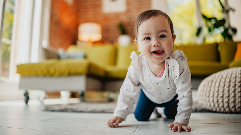 A baby girl crawls in a living room with a happy expression and her tongue sticking out