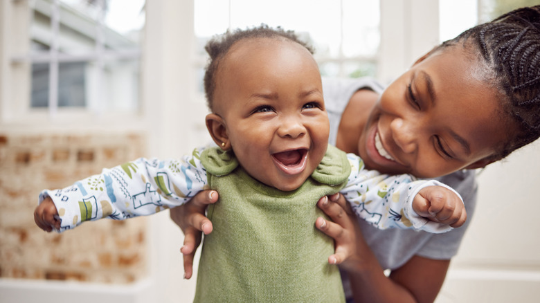 A mother holds a baby girl up underneath the armpits, both with happy and cute expressions on their faces