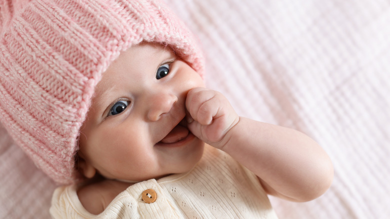 A baby girl wearing a pink winter hat smiles with her tongue sticking out, eyes wide open, and a thumb in her mouth