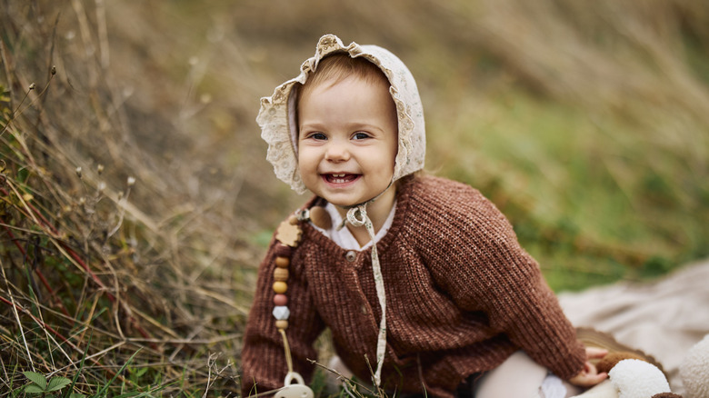 A toddler girl wearing a sweater and bonnet smiles while sitting in nature