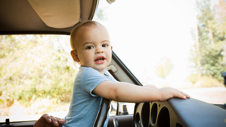A baby boy leans on the dash board inside a vehicle with a smiling expression