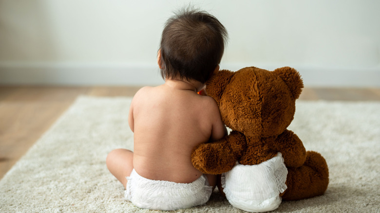 A baby and a teddy bear, both wearing diapers, sit beside each other in a side hug