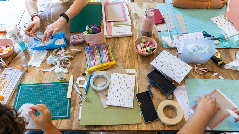 A craft table covered with supplies with peoples' arms in view