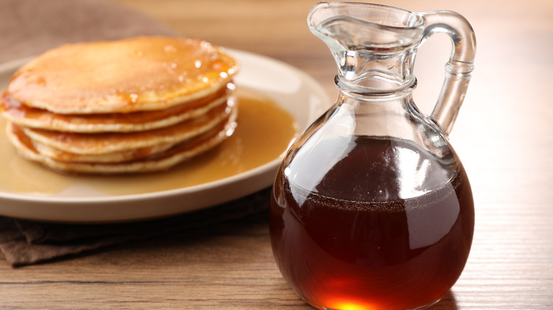 A glass container of pure maple syrup with a stack of pancakes in the background