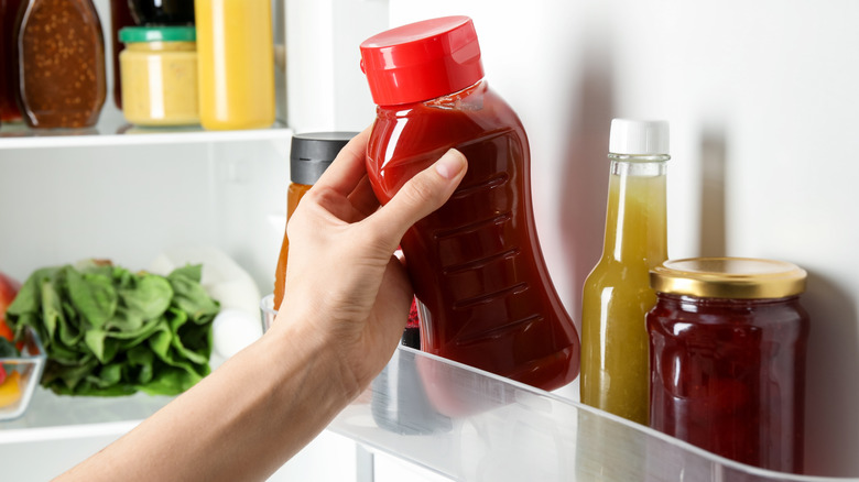 A person reaches for ketchup from the condiments shelf in a refrigerator