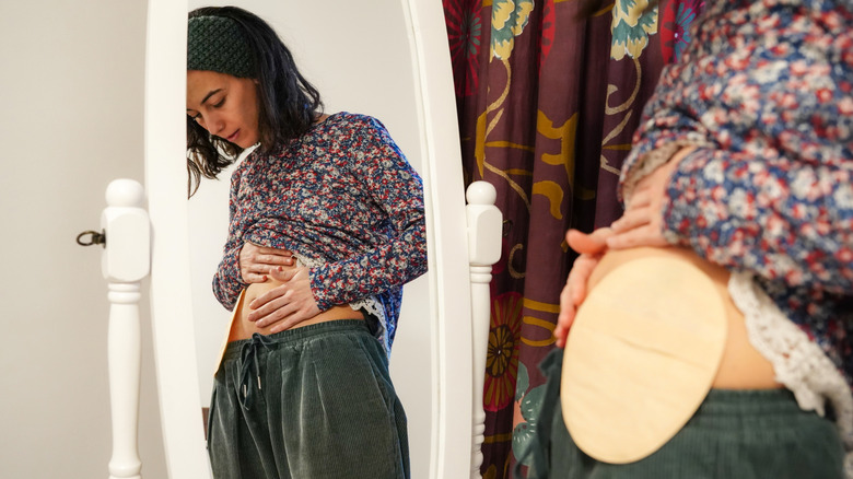 A woman pulling up shirt and touching stomach near ostomy bag in front of the mirror