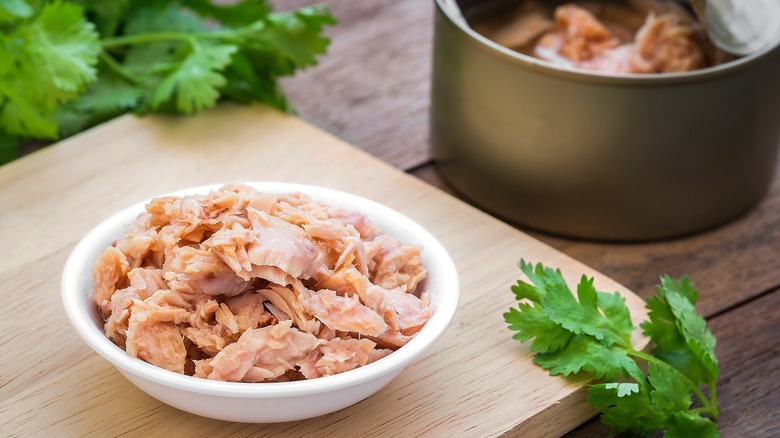 Canned tuna in a small white bowl on a wooden serving board surrounded by fresh cilantro with a can of opened tuna in the background