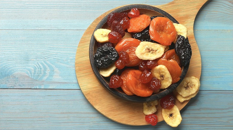 Bowl of dried apricots, plums, cranberries, and banana chips on a round wooden serving board over a light blue background