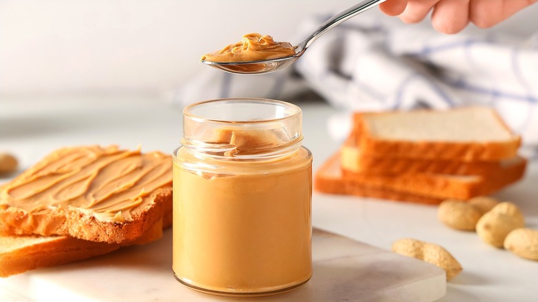A hand holding a metal spoon full of peanut butter over a peanut butter jar surrounded by slices of bread and whole, loose peanuts