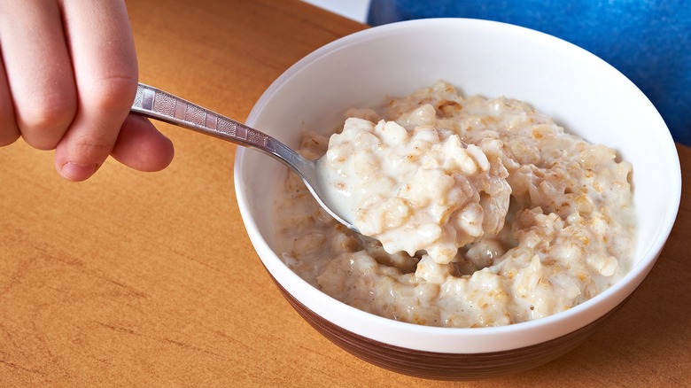 A child's hand holding a metal spoon in a bowl of creamy oatmeal
