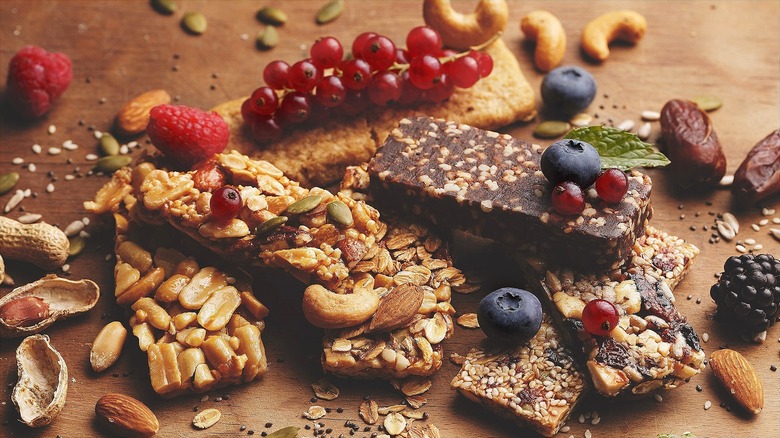 A variety of whole-food snack bars surrounded by fresh and dried fruit and nuts on a wooden surface