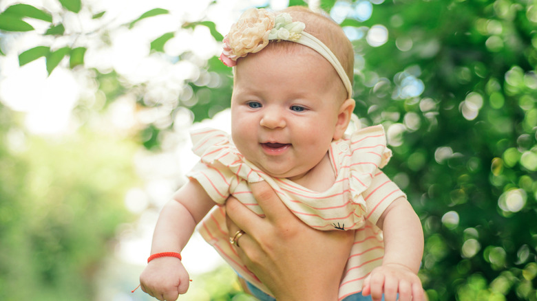 A baby girl with a flower headband is held up in front of lush greenery outside