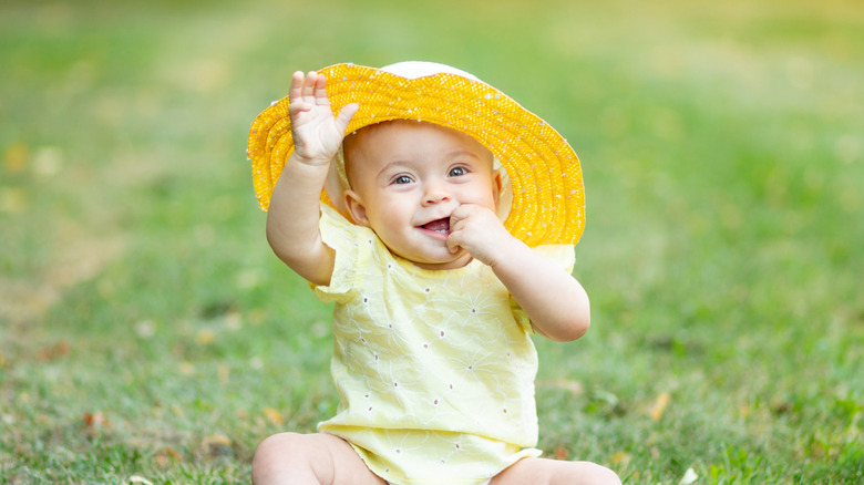 A baby girl wears a sun hat and waves at the camera, sitting outside on the grass