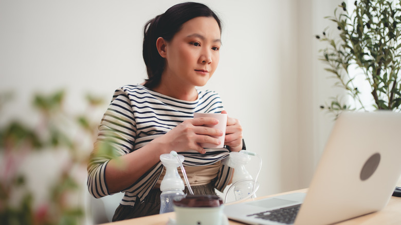 woman working at laptop and drinking while using electric breast pump