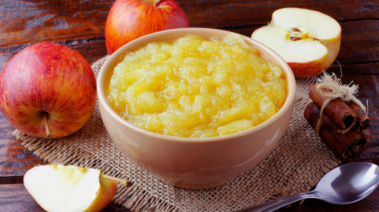 Bowl of applesauce sitting on a woven placemat with apples, a stack of cinnamon, and a metal spoon