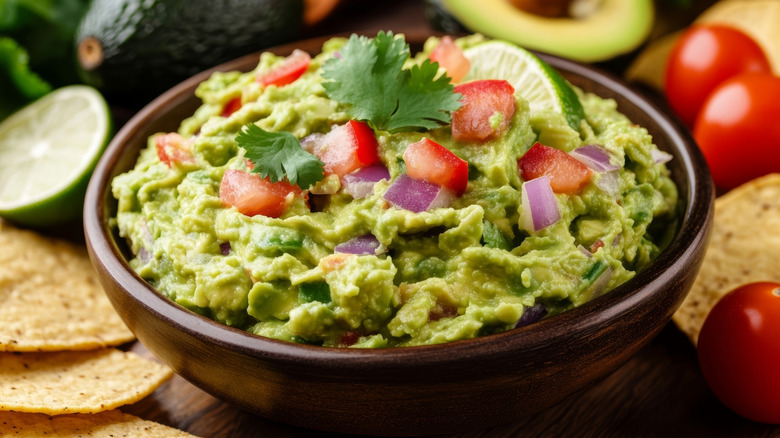 Wooden bowl filled with guacamole with chips, tomatoes, avocados, and a lime