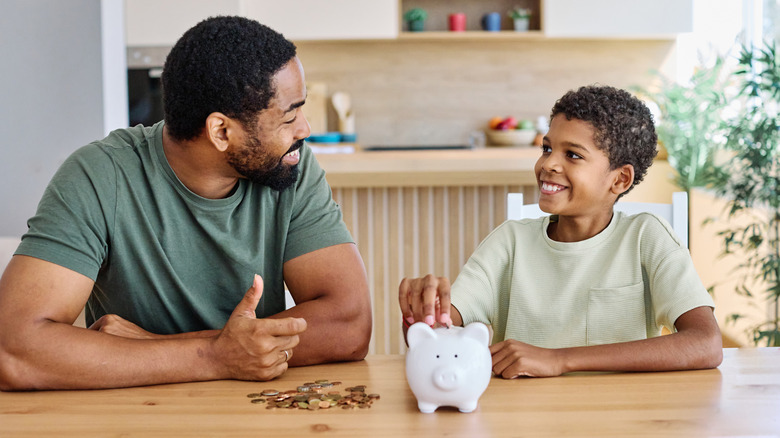 A young boy smiles at an adult male while placing his hand on a piggy bank with a pile of coins on the table