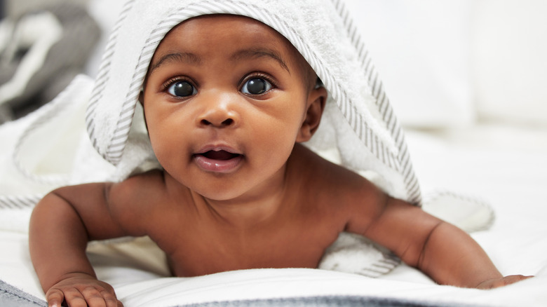 A baby holds herself on her stomach up wearing a hooded towel and looks right at the camera