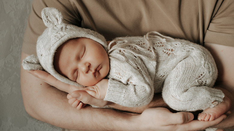 An infant sleeps in her parent's arms dressed in a soft knitted bunny outfit