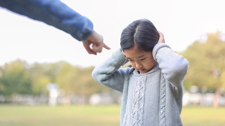 A child holds her hands to her ears and looks down at the grass while a finger points in her face