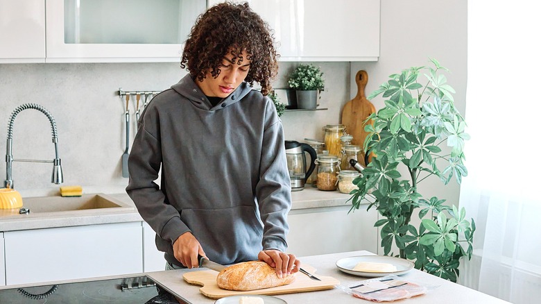 Teenage boy with dark, curly hair cutting a loaf of bread in a white and gray kitchen