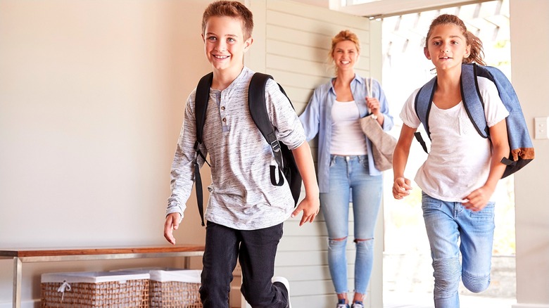 Two kids in backpacks returning home after school with a woman wearing jeans smiling behind them