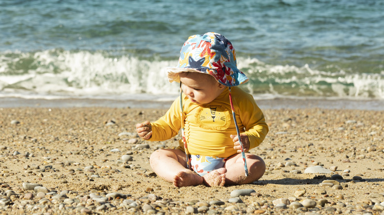 baby playing on the sand at the beach wearing a hat and rash guard