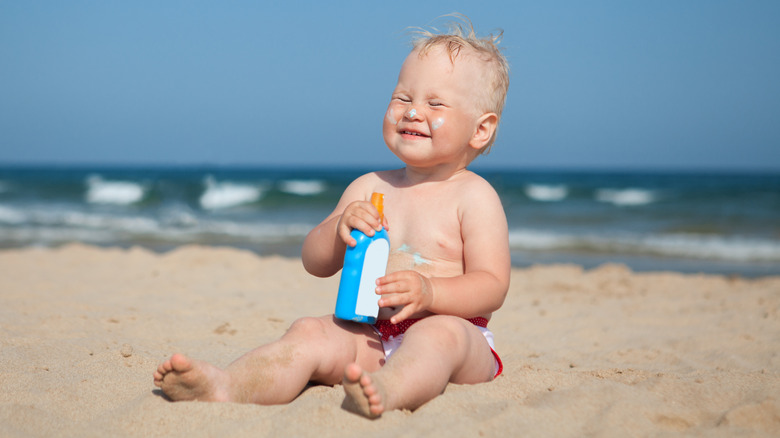 baby sitting on the sand with eyes closed, holding a bottle of sunscreen and wearing sunscreen on nose and cheeks