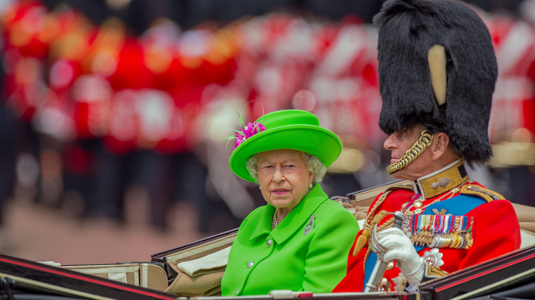 Queen Elizabeth II of England riding in a car with a royal guard wearing a green jacket and hat