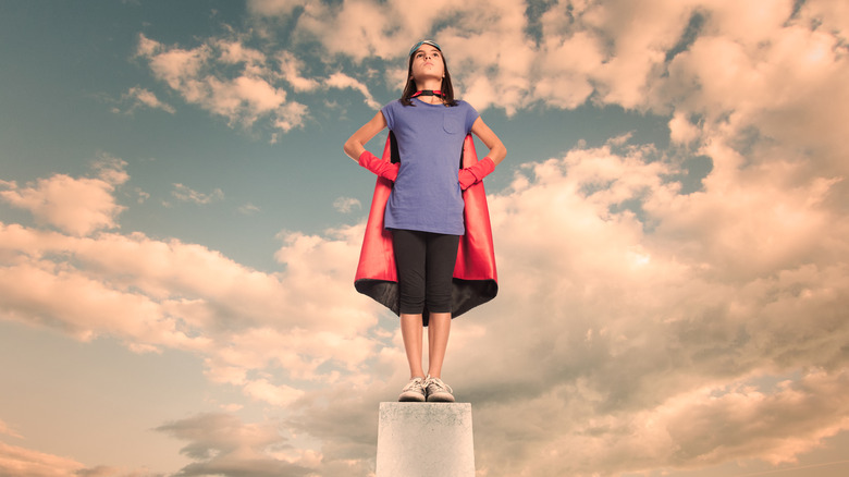 Young girl standing on top of a pillar wearing a heroe's cape with the sky in the background