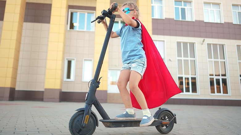 Child wearing a red cape and an eye mask while riding an electronic scooter in front of a large building