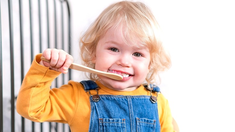 Toddler with shaggy blonde hair brushing teeth, wearing a yellow long sleeve shirt and overalls