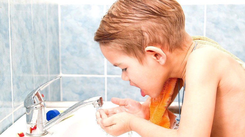 Shirtless toddler boy rinsing his mouth over a sink with both hands