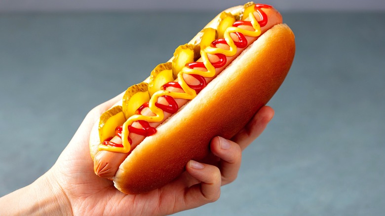 A hand holding a hot dog in a bun with ketchup, mustard, and pickle slices over a gray background