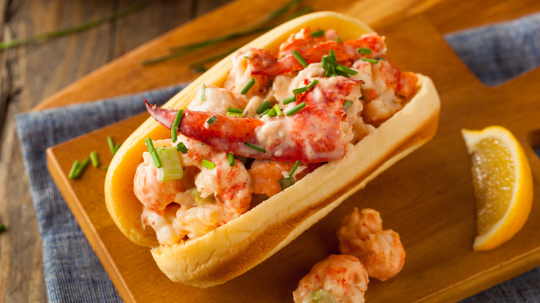 A cooked lobster roll with garnishes sits on top of a wooden cutting board